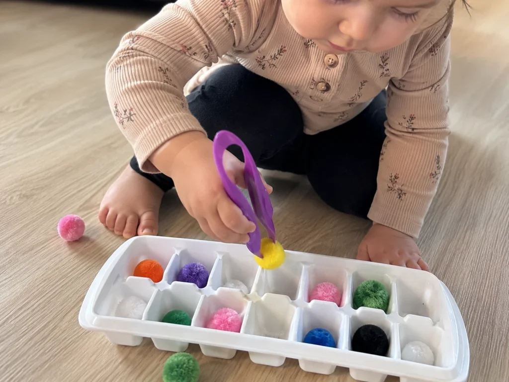 toddler using tongs to sort colored pom-poms into an ice cube tray